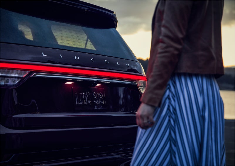 A person is shown near the rear of a 2023 Lincoln Aviator® SUV as the Lincoln Embrace illuminates the rear lights | Boulevard Lincoln in Millsboro DE